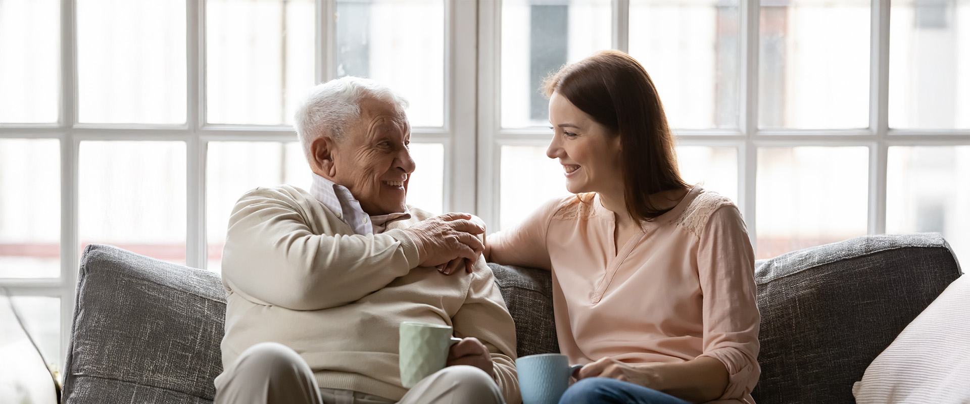 Elderly man and young woman smiling while sitting on a sofa, holding mugs